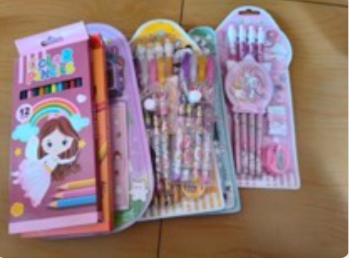A child's stationery set on a wooden floor: a twelve-pack of colouring pencils, gel pens, and unicorn-themed pencil and eraser sets, all in their retail packaging.