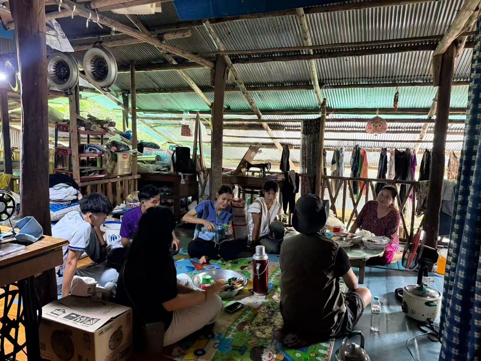 Seven people sit on the floor under a corrugated-metal roof, most of them on a patterned mat. A young woman pours water from a bottle while an older woman sits by a low table of dishes, with sewing machines and hanging clothes around the room.