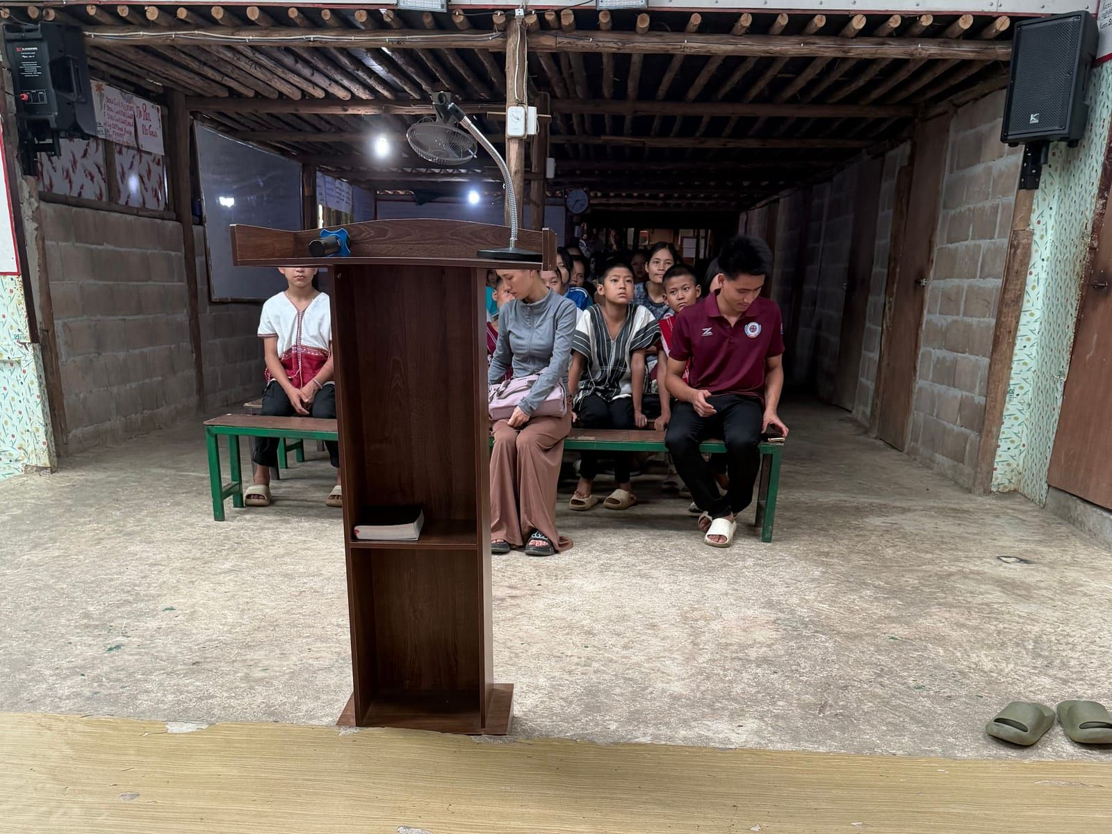 A wooden lectern with a microphone stands in front of green benches where children and a few adults sit facing the camera, in a hall with breeze-block walls and a timber ceiling.
