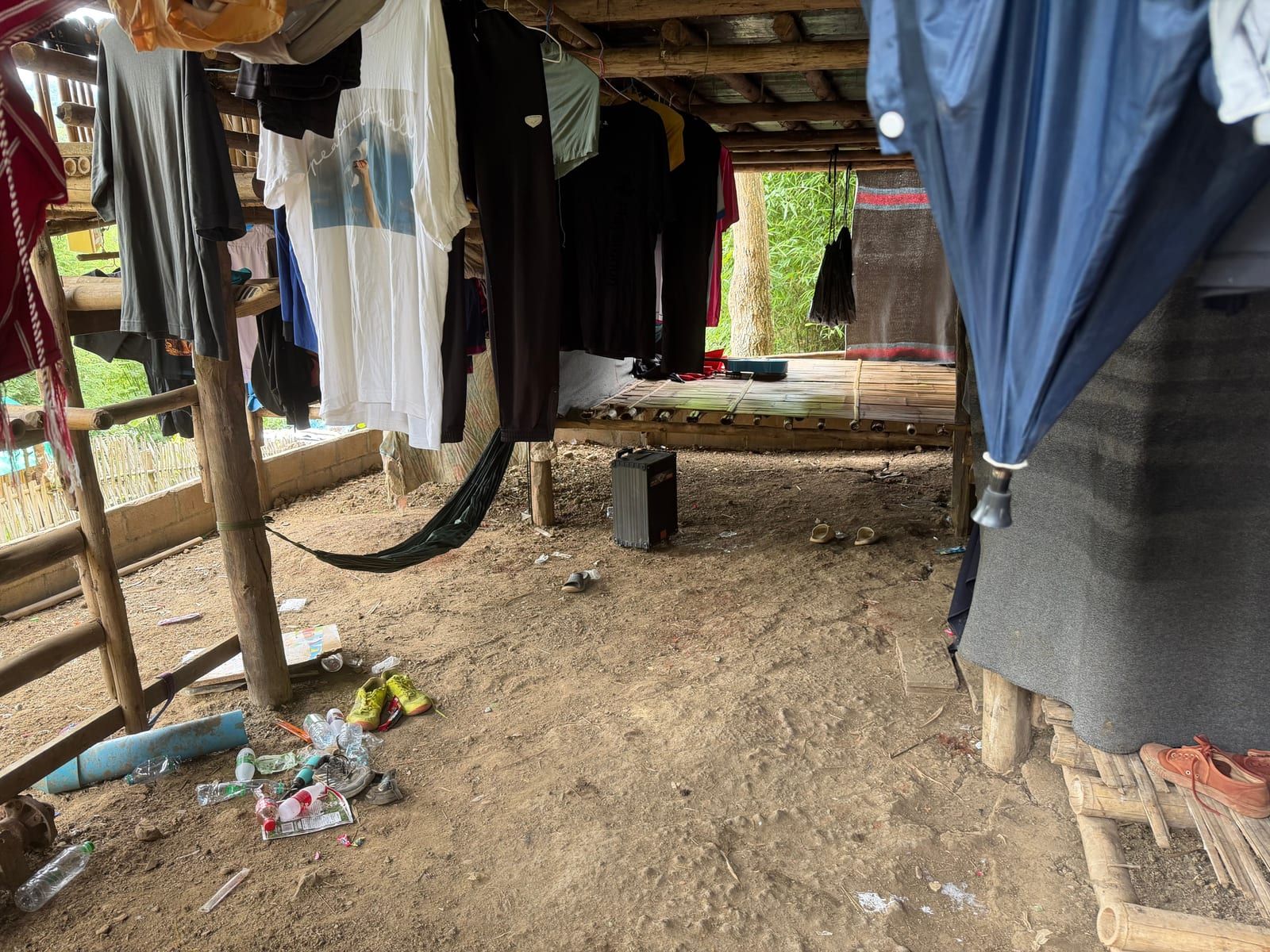 The shaded space beneath a raised house: a packed-dirt floor, wooden posts, rows of shirts on hangers, a hammock, a loudspeaker and a low bamboo platform at the back.