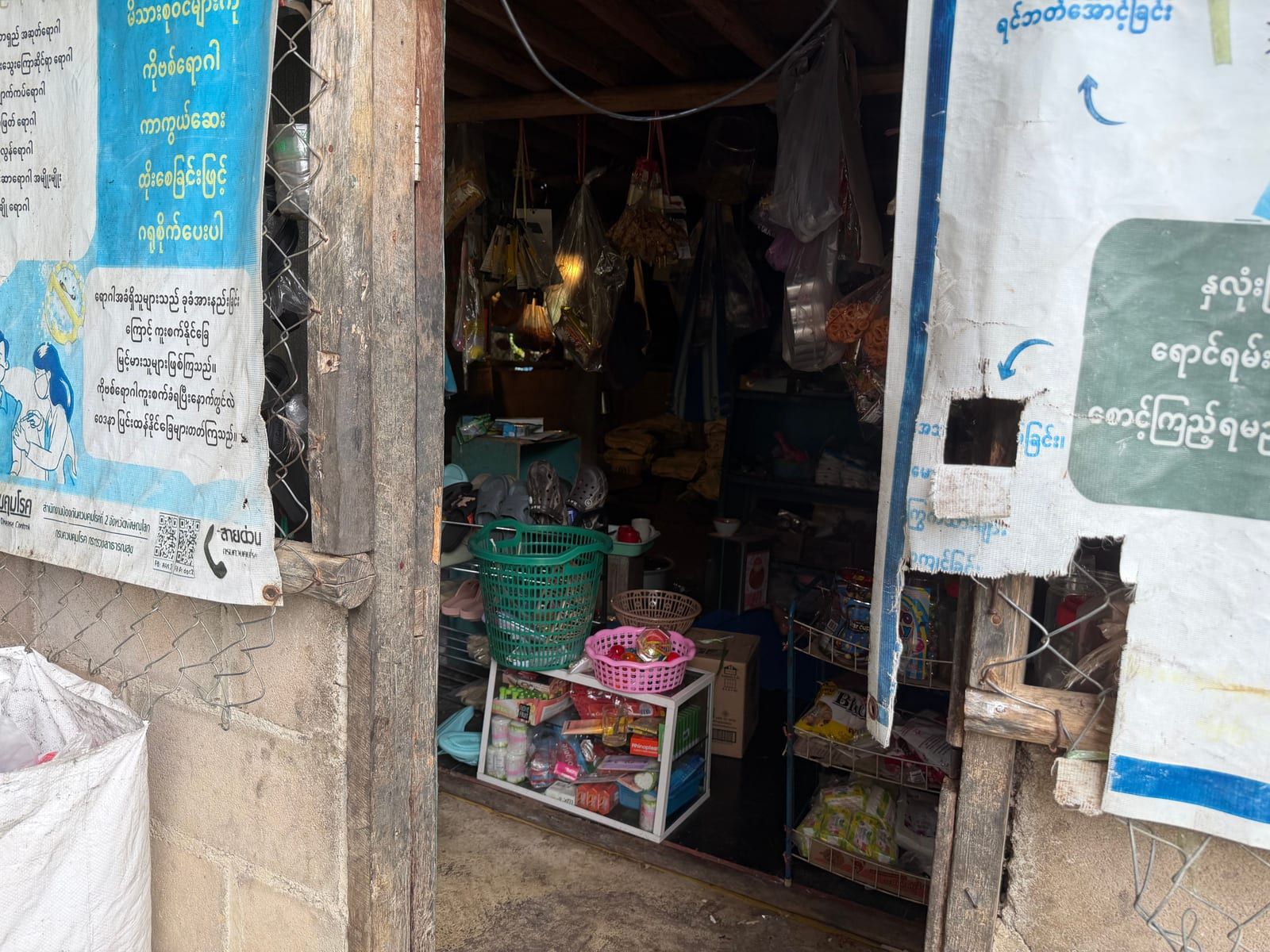 A narrow shop doorway between two weathered posters in Burmese script. Inside, bagged snacks hang from the ceiling above baskets, a shoe rack and shelves of packaged goods.