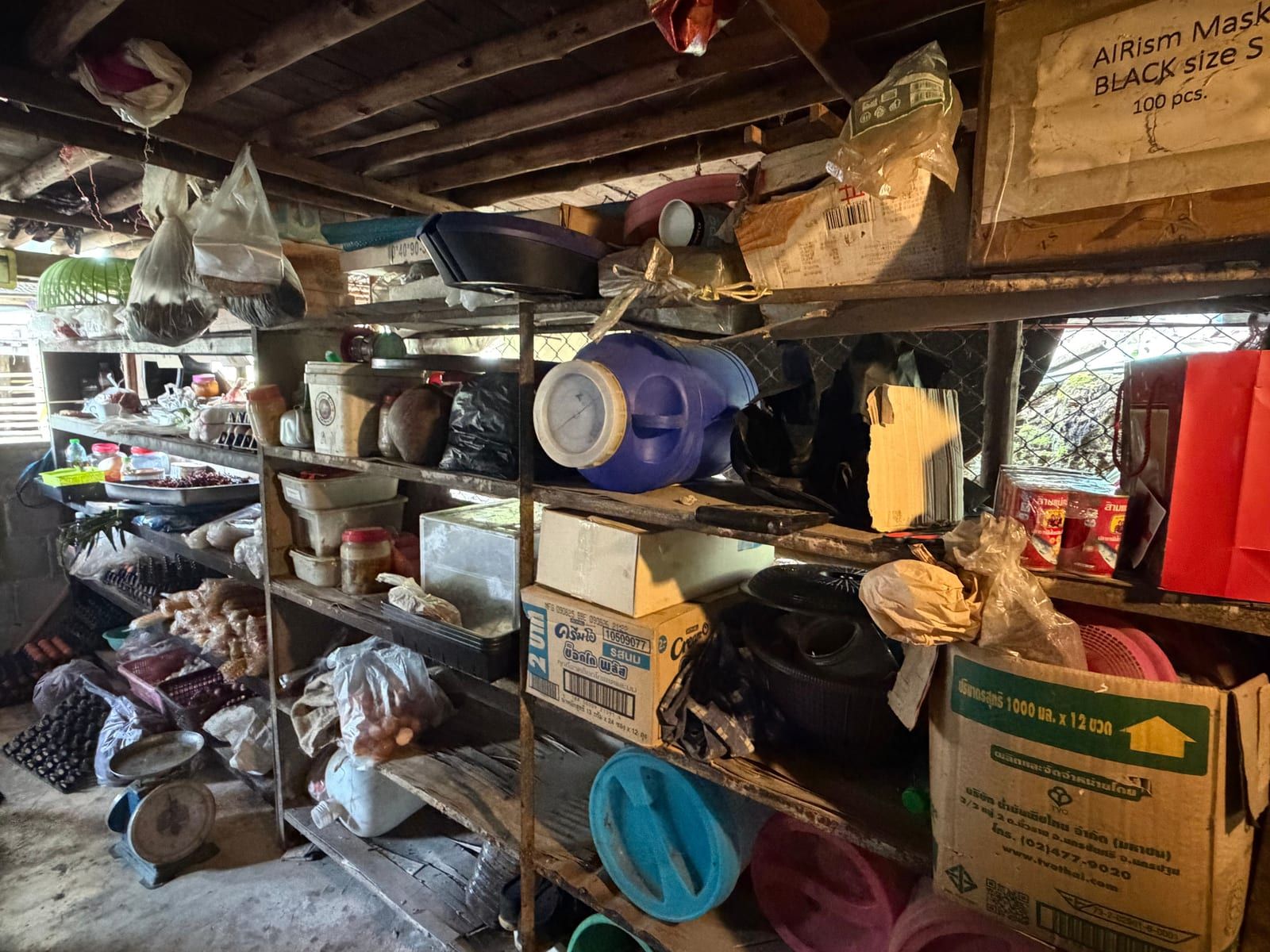 A dim storeroom lined with shelves holding cardboard boxes, plastic tubs, a blue plastic drum, jars and egg trays, with a dial weighing scale on the concrete floor.