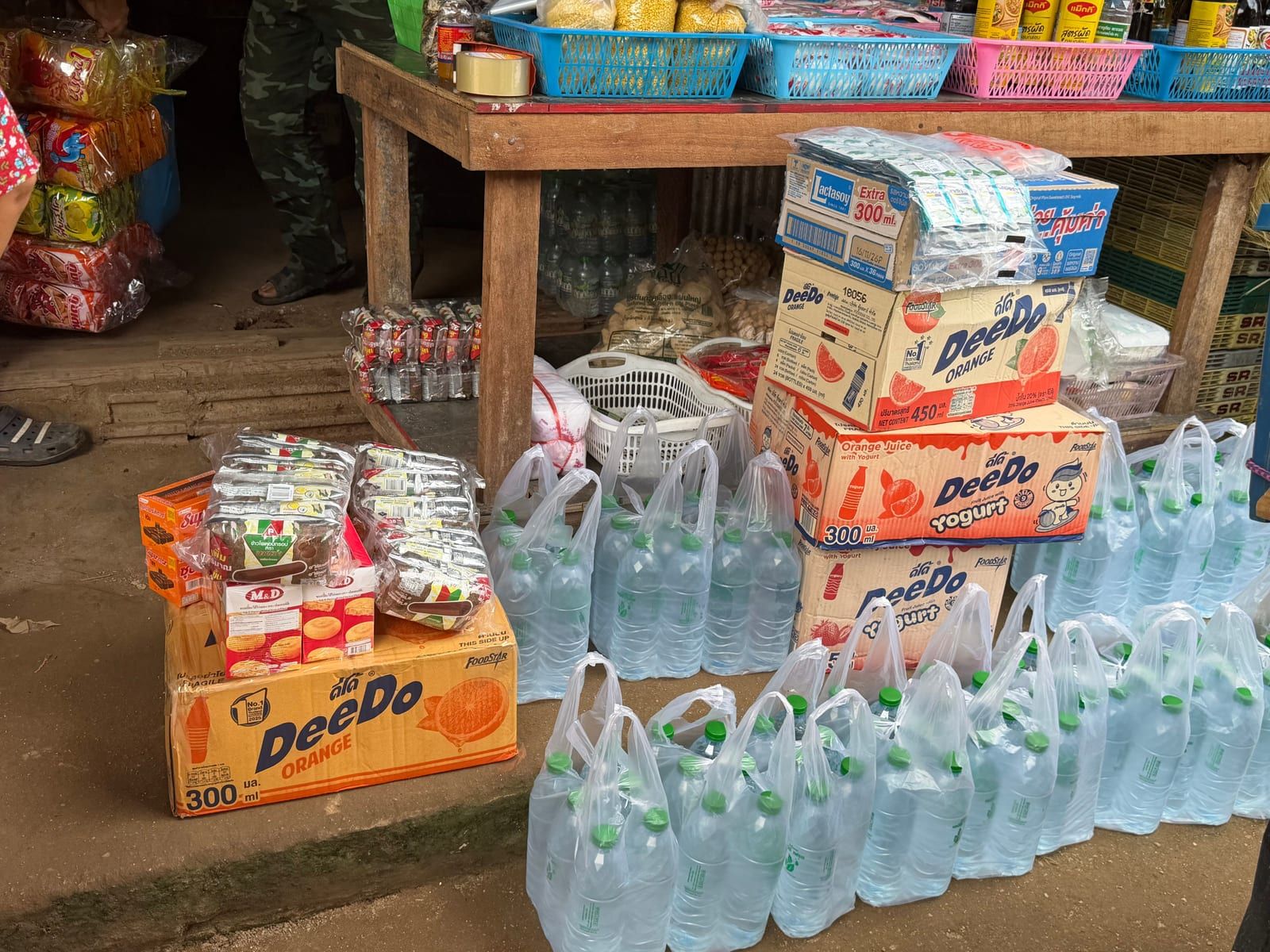 Cardboard cases of orange juice, packs of biscuits and rows of plastic bags holding bottled water, stacked on the ground in front of a wooden table of baskets and packaged food.